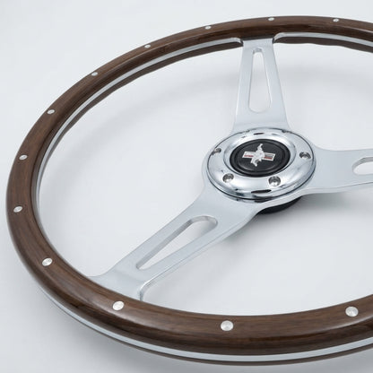 A vintage-style steering wheel with a dark brown wood rim, polished silver metal spokes, and a central chrome hub featuring a black Mustang logo.