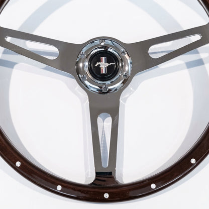 A close-up of a vintage-style steering wheel with a dark brown wood rim, polished silver metal spokes, and a chrome center cap featuring a Mustang logo.
