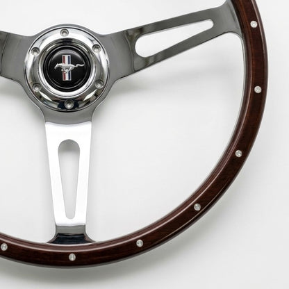 A close-up of a vintage-style Ford steering wheel featuring a dark brown wood rim with silver rivets, polished chrome spokes, and a central Mustang logo badge.