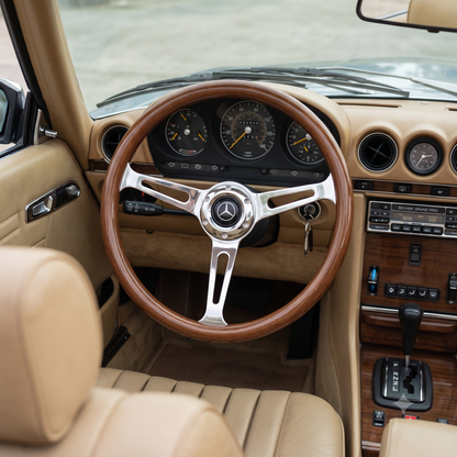 A close-up view of a classic Mercedes-Benz car interior, featuring a wood-rimmed steering wheel with a polished chrome center and a beige leather dashboard and seats.