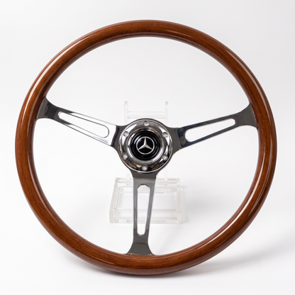 A vintage Mercedes-Benz steering wheel with a polished brown wood rim and polished silver metal spokes, featuring the Mercedes-Benz star logo in the center, displayed on a clear stand against a white background.