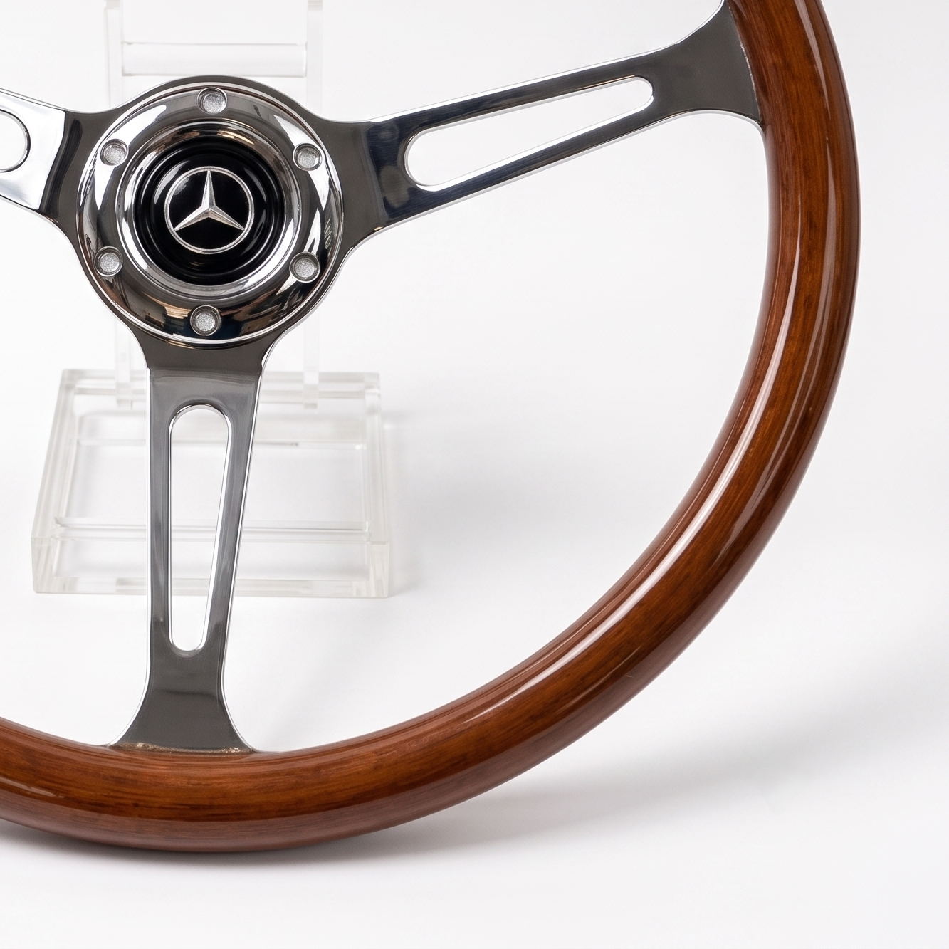 A close-up of a Mercedes-Benz steering wheel with a polished brown wood rim and polished silver metal spokes, displayed on a clear stand against a white background.