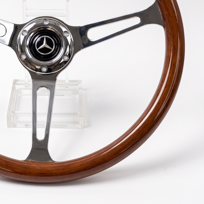 A close-up of a Mercedes-Benz steering wheel with a polished brown wood rim and polished silver metal spokes, displayed on a clear stand against a white background.