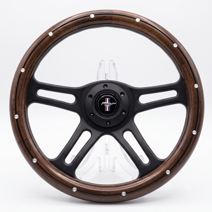 A custom four-spoke steering wheel featuring a dark brown wood grain outer rim, a matte black center and spokes, and silver rivet accents around the outer edge. The center cap displays a Mustang logo.