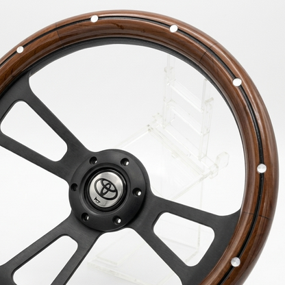 A close-up of a car steering wheel featuring a polished brown wood rim, black multi-spoke center, and silver Toyota emblem in the hub.